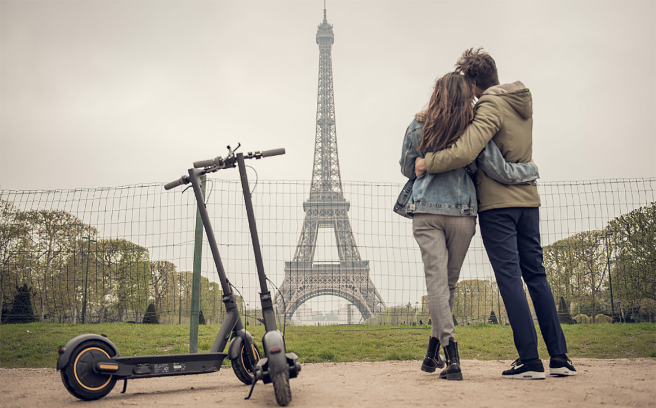 A view from behind a couple staring at a landmark embracing with two Segway Ninebot G30P Max electric scooters off to the side.